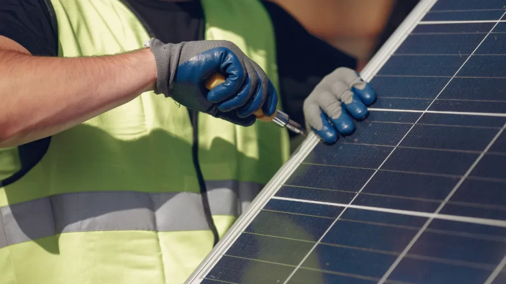 Técnico realizando manutenção em placa solar com ferramenta manual, utilizando luvas de proteção e colete de segurança.
