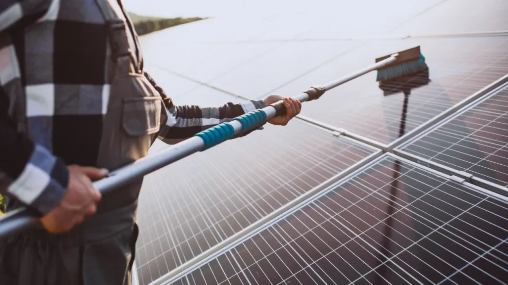 Pessoa realizando limpeza de placas solares com escova telescópica em telhado, removendo sujeira do painel fotovoltaico.