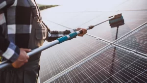 Pessoa realizando limpeza de placas solares com escova telescópica em telhado, removendo sujeira do painel fotovoltaico.